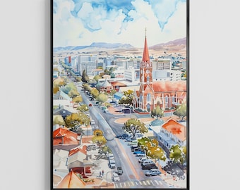 May include: Watercolor painting of a city street with a church and buildings. The artwork features a red-roofed church with a tall steeple, a road lined with trees, and a blue sky with clouds. The painting is framed in a black frame.