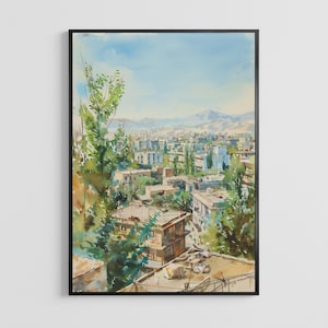 May include: Watercolor painting of a cityscape with buildings, trees, and mountains in the background. The artwork is framed in a black frame. The color palette includes shades of blue, green, and brown.