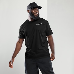 May include: Black athletic t-shirt with the Greek word "υπομονή" embroidered in white. The shirt is short-sleeved and has a crew neck. The person in the image is wearing a black trucker hat and black pants.