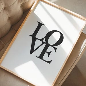 Könnte beinhalten: Ein gerahmter Kunstdruck mit dem Wort "LOVE" in fetten schwarzen Buchstaben. Der Druck hat einen weißen Hintergrund und einen hellbraunen Holzrahmen. Das Kunstwerk liegt auf einem beigefarbenen Polstersessel.