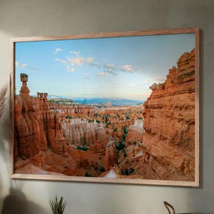 Puede incluir: Una fotografía enmarcada de un paisaje de cañón con formaciones rocosas anaranjadas bajo un cielo azul con nubes. La fotografía se exhibe sobre un mueble de madera con objetos decorativos, que incluyen un jarrón con plantas secas, una pequeña planta en maceta, una vela y una cesta tejida.