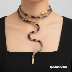 May include: A snake-shaped necklace with a gold-coloured head and tail. The necklace is wrapped around the neck and drapes down the chest. The snake's body is a mix of black, tan, and orange colours. The necklace is worn over a black top.