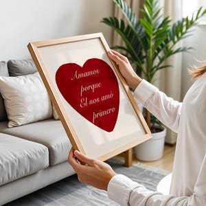 May include: A framed print featuring a large red heart with the Spanish text "Amamos porque El nos am&oacute; primero" in a light-colored font. The frame is made of light-colored wood.