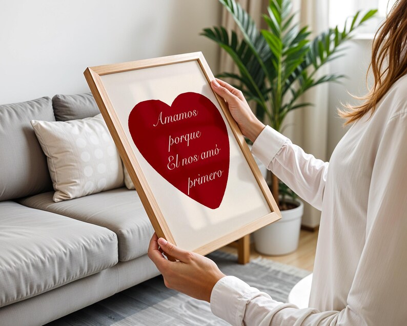 May include: A framed print featuring a large red heart with the Spanish text "Amamos porque El nos am&oacute; primero" in a light-colored font. The frame is made of light-colored wood.