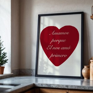 May include: A framed print featuring a large red heart on a white background. The heart has the words "Amamos porque El nos am&oacute; primero" in a script font. The frame is black and the print is displayed on a kitchen counter.