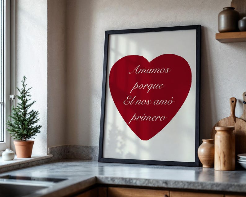 May include: A framed print featuring a large red heart on a white background. The heart has the words "Amamos porque El nos am&oacute; primero" in a script font. The frame is black and the print is displayed on a kitchen counter.