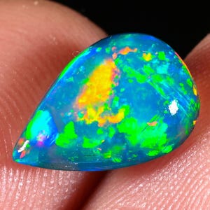 May include: A pear-shaped opal gemstone with a vibrant display of colors. The stone features a base of deep blue, with flashes of green, yellow, and orange. The opal is held between fingers against a dark background.