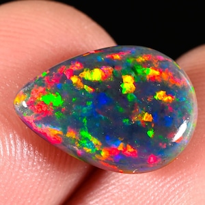 Può includere: Una pietra preziosa di opale nero a forma di goccia con vibranti lampi di colore, tra cui rosso, verde, giallo e blu. La superficie lucida riflette la luce, mostrando il gioco di colori interno. La pietra preziosa è tenuta tra due dita.