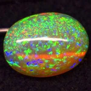 May include: A vibrant, oval-shaped opal gemstone. The opal displays a spectrum of colors, including green, orange, blue, and yellow, with a bright, iridescent play of color. The gemstone is set against a dark background.