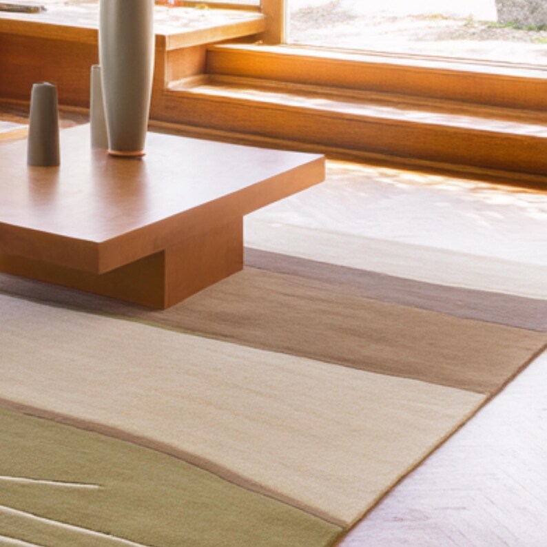 May include: A rectangular area rug with a striped pattern in shades of green, beige, and brown. The rug is placed on a light-colored wooden floor, with a wooden coffee table and decorative vases in the background.