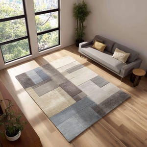 Puede incluir: Una alfombra rectangular con un diseño geométrico en tonos beige, gris y azul. La alfombra está colocada sobre un suelo de madera, con un sofá gris y una pequeña mesa auxiliar en el fondo. Se ve una ventana grande.