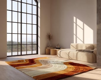 May include: A rectangular area rug with a wavy, striped design in shades of orange, brown, and cream. The rug is placed on a wooden floor in a well-lit room with a large window and a beige sofa. The text "Rug America" is visible on the rug.