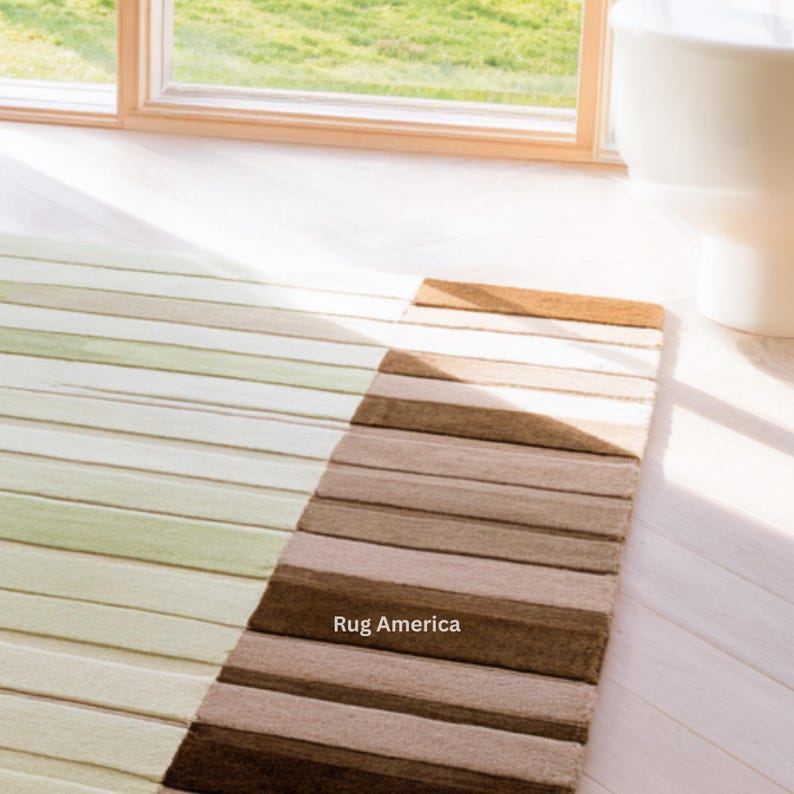 Pu&ograve; includere: Un tappeto rettangolare a righe orizzontali nei toni del verde, beige e marrone. Il tappeto &egrave; posizionato su un pavimento in legno bianco vicino a una finestra. Il testo "Rug America" &egrave; visibile sul tappeto.