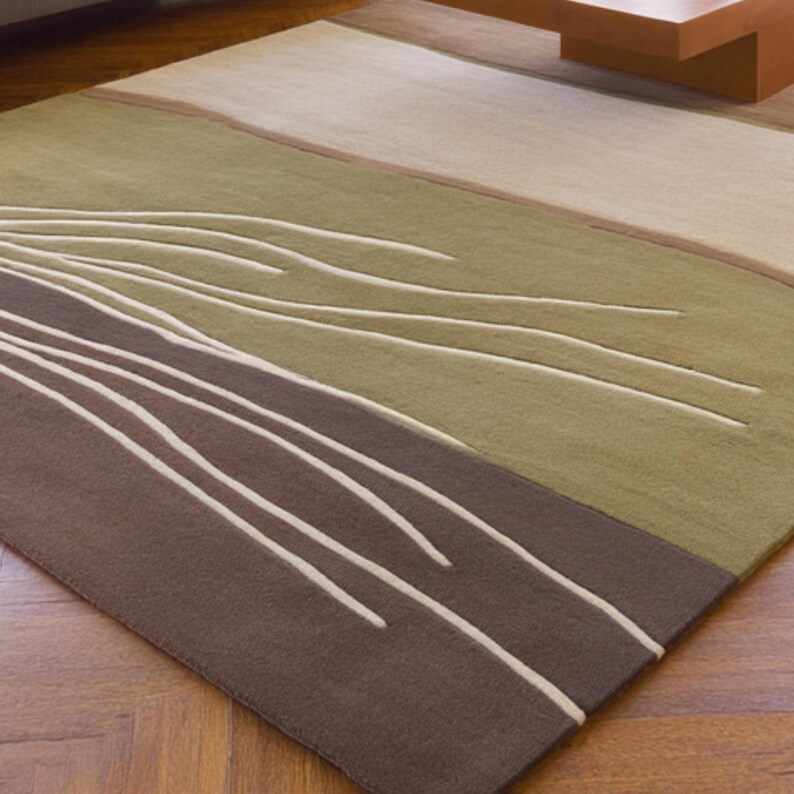 May include: A rectangular area rug with a modern design. The rug features a color block pattern with shades of brown, green, and beige. White, curved lines create a flowing design across the rug's surface. The rug is made of a soft, textured material.