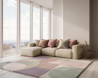 May include: A beige sectional sofa with several decorative pillows in shades of pink, green, and beige. A large area rug with a geometric pattern in pastel colors lies on the wooden floor. The rug has the text "Rug America".