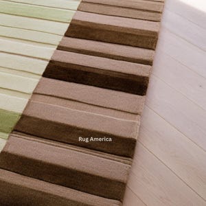 Pu&ograve; includere: Un tappeto rettangolare a righe orizzontali nei toni del verde, marrone e beige. Il tappeto &egrave; realizzato in un materiale strutturato ed &egrave; posizionato su un pavimento in legno chiaro. Il testo "Rug America" &egrave; visibile sul tappeto.