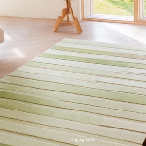 Pu&ograve; includere: Un tappeto rettangolare con un motivo a righe in tonalit&agrave; di verde e beige. Il tappeto &egrave; realizzato in un materiale morbido e ha un aspetto strutturato. Il tappeto &egrave; posizionato su un pavimento in legno chiaro. Il testo "Rug America" &egrave; in basso.