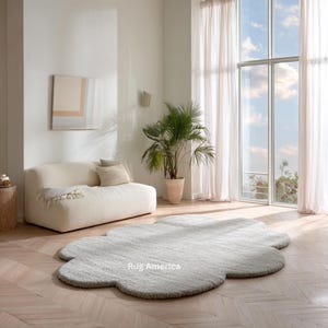 May include: A pale grey, cloud-shaped rug sits on a light wood floor in a bright room. The rug has a soft, textured appearance. A white sofa, a potted plant, and a large window with sheer curtains are also visible.