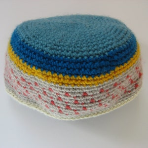 May include: A handmade, crocheted hat with a rounded crown. The hat features a light blue top, a blue band, a yellow band, and a speckled gray and red lower section. The brim is a cream color. The hat is made of yarn.