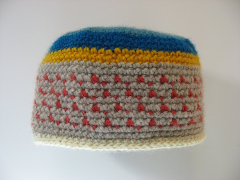 May include: A handmade crochet beanie in a color block design. The hat features a light gray base with red accents, a yellow stripe, and a blue stripe at the top. The brim is a cream color.