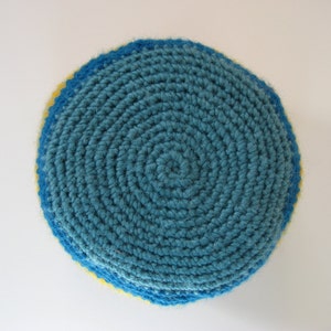 May include: A handmade, circular crochet item in shades of blue and yellow. The center is a teal blue, spiraling outward. The edges are trimmed with a darker blue, and a thin yellow band is visible. The item is likely a coaster or trivet.