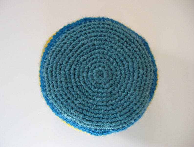 May include: A handmade, circular crochet item in shades of blue and yellow. The center is a teal blue, spiraling outward. The edges are trimmed with a darker blue, and a thin yellow band is visible. The item is likely a coaster or trivet.