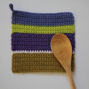 May include: A handmade, square crochet potholder with a loop for hanging. The potholder features horizontal stripes of blue, green, purple, white, and olive green. A wooden spoon rests on the potholder.