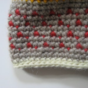 May include: Close-up of a crocheted item with a textured pattern. The main color is a light gray, with small red dots evenly spaced throughout. A band of cream-colored yarn forms the bottom edge, and a yellow band is visible at the top.
