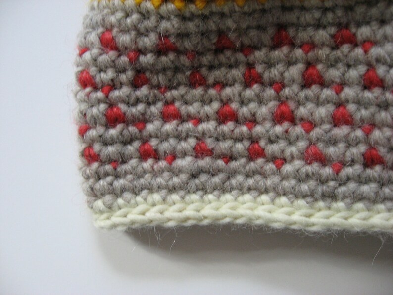 May include: Close-up of a crocheted item with a textured pattern. The main color is a light gray, with small red dots evenly spaced throughout. A band of cream-colored yarn forms the bottom edge, and a yellow band is visible at the top.