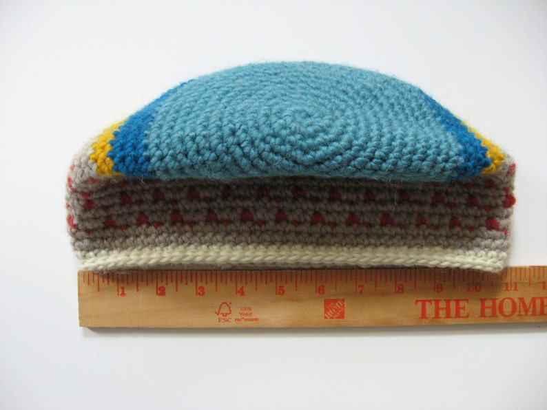 May include: A handmade crochet hat with a blue top, a yellow and blue stripe, and a gray and red patterned band. The hat measures approximately 11 inches in length.