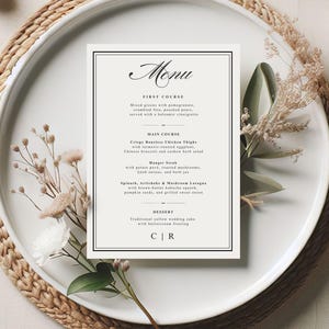 May include: A formal menu card with a black border and elegant script font, featuring a list of dishes. The card is placed on a white plate with decorative floral elements. The menu includes first course, main course, and dessert options.