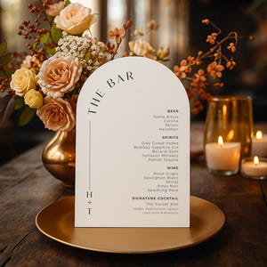 May include: A white, arched bar menu displaying a drinks list, including beer, spirits, wine, and a signature cocktail. The menu sits on a gold plate, with flowers and candles in the background. The text "THE BAR" is at the top.