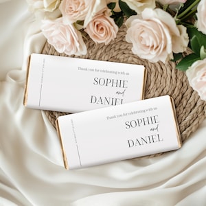 Könnte beinhalten: Zwei Schokoriegel mit weißen Verpackungen, bedruckt mit "SOPHIE und DANIEL" in schwarzer, eleganter Schrift. Die Verpackungen enthalten auch den Text "Vielen Dank, dass Sie mit uns feiern" und ein Datum. Die Riegel liegen auf einer gewebten Matte, mit rosa Rosen im Hintergrund.