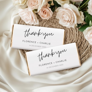 Könnte beinhalten: Zwei weiße Schokoladenriegelverpackungen mit dem handschriftlichen Schriftzug "thank you". Die Namen "Florence + Charlie" und das Datum "14. Dezember 2025" sind darunter aufgedruckt. Die Riegel liegen auf einer geflochtenen Matte mit Blumen.