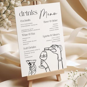 May include: A drinks menu displayed on a small wooden easel. The menu, printed in black script, lists cocktails, beer & cider, wine & fizz, spirits & mixers, and soft drinks. The design includes line art illustrations of hands pouring drinks.