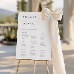 Peut inclure: Un plan de table de mariage blanc sur un chevalet doré, avec les noms "Shelby et Devon" et la date "Samedi 24 septembre 2025." Un grand nœud crème orne le coin supérieur droit. Les noms des invités sont listés par table.