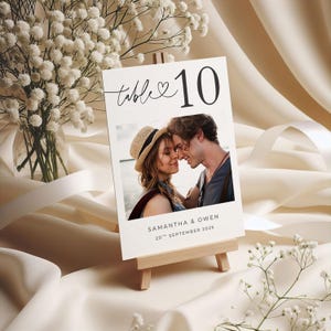 Puede incluir: Una tarjeta con el número de mesa de boda sobre un pequeño caballete de madera. La tarjeta presenta el número 10 en negro, con la palabra "mesa" en cursiva. Una foto de una pareja está debajo, con los nombres "Samantha & Owen" y la fecha.