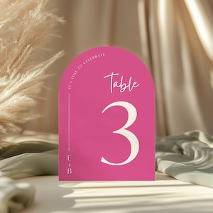 May include: A hot pink table number card with an arched top. The card features the word "Table" in a script font, the number "3" in a bold font, and the text "It's time to celebrate". The card also has the initials "E + B".