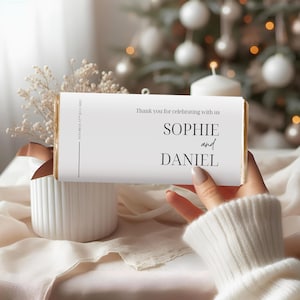 Op de afbeelding: Een witte chocoladereep met gouden randen, met de tekst "Thank you for celebrating with us" en de namen "SOPHIE and DANIEL". De reep wordt in een hand gehouden, met een vage kerstboom op de achtergrond.