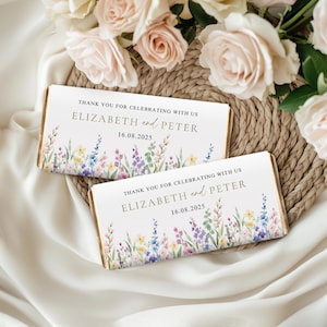 May include: Two rectangular wedding favours with floral watercolour designs. The favours have the text "THANK YOU FOR CELEBRATING WITH US" and the names "ELIZABETH and PETER" with the date "16.08.2025". The favours are on a woven mat with flowers in the background.