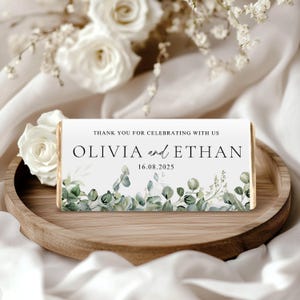 May include: A rectangular chocolate bar wrapper with the text "THANK YOU FOR CELEBRATING WITH US" and the names "OLIVIA and ETHAN" along with the date "16.08.2025". The design includes green eucalyptus leaves and floral accents.
