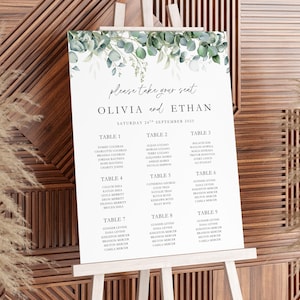 Può includere: Un tableau di matrimonio bianco su un cavalletto, adornato con verde eucalipto. Il grafico mostra i nomi di Olivia ed Ethan, insieme ai posti a sedere degli ospiti per i tavoli 1-9. La data è sabato 24 settembre 2025.
