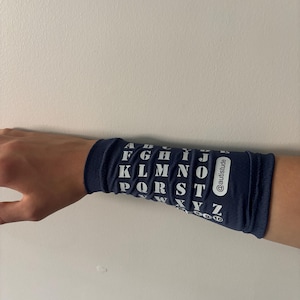 May include: A navy blue wristband featuring the alphabet in white block letters. The wristband also has the text "@autistude" printed on it. The wristband is worn on a person's wrist.