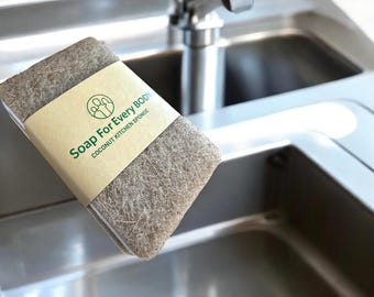 May include: A rectangular, gray coconut kitchen sponge with a brown paper label wrapped around it. The label features the text "Soap For Every Body" and "Coconut Kitchen Sponge" in green. The sponge is positioned near a stainless steel sink and faucet.