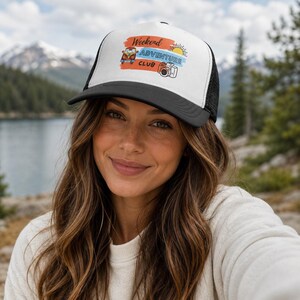 May include: A black and white trucker hat with the words "Weekend Adventure Club" in orange and blue. The hat features a graphic of a camper van, a camera, and a sun. The hat is worn by a person with long brown hair.
