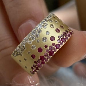 May include: A gold-toned ring with a band design. The ring is adorned with a gradient of gemstones, including clear and ruby-colored stones. The gemstones are set in a pattern of varying sizes, creating a textured appearance. The ring is held by a person.
