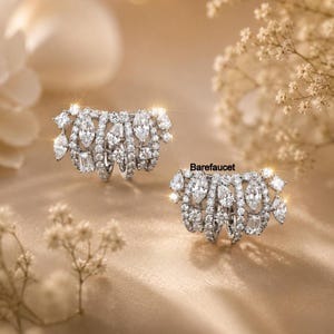 May include: A pair of silver-toned ear cuffs featuring multiple rows of clear, faceted stones in marquise, round, and baguette cuts. The text Barefaucet is printed near the right ear cuff. These jewelry pieces have a metallic finish.