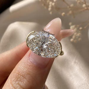 Puede incluir: Un anillo con un diamante de corte ovalado que presenta una piedra grande, transparente y facetada, montada en una banda de oro con engastes laterales. El texto Barefaucet está impreso en la superficie del diamante.
