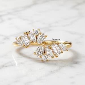 May include: A gold ring with a cluster of diamonds. The ring features a central emerald-cut diamond flanked by pear-shaped and round diamonds. The ring is set against a white marble surface. The text "Barefaucet" is visible.