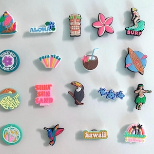 May include: A collection of colorful tropical-themed charms. Designs include a flamingo, tiki, flower, surfer, coconut drink, toucan, and phrases like "Aloha" and "Hawaii." These charms are likely for shoes or accessories.